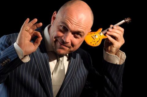 Wolf Martini hält sich eine Minigitarre ans linke Ohr und macht mit der rechten Hand ein OK-Zeichen.